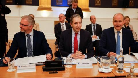 Taoiseach and Fine Gael leader Simon Harris (middle) , Fianna Fáil leader Micheál Martin (right) and Green Party leader Roderic O'Gorman (left) sit at a table with name tags in front of them. Simon Harris is wearing a bright red tie and dark blazer, Roderic is wearing a black and white speckled tie and wears glasses. Micheál is wearing a light blue tie and navy blazer.