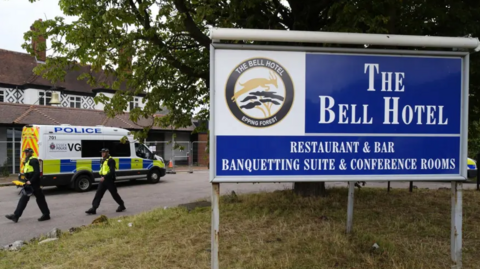 A large blue and white sign in the grounds outside The Bell Hotel in Epping. In the background there are two police officers in hi-vis jackets and a police van.