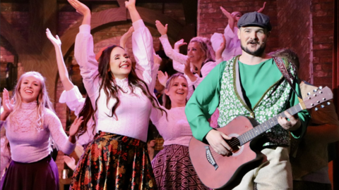 A man plays a guitar and sings while a group of women in skirts and white tops dance around him. He wears a flat cap and a shamrock pattern waistcoat.