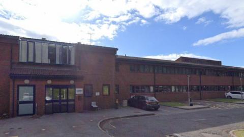 Two storey red brick building with lots of windows to the right of the picture and a set of doors to the left of the building