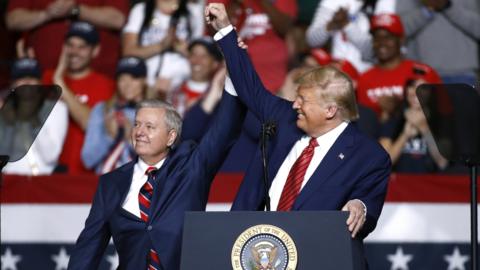 President Donald Trump and Senator Lindsey Graham are wearing navy blue business suits and standing behind a podium with the US presidential seal holding, each holding an arm up toward the other in a victorious gesture