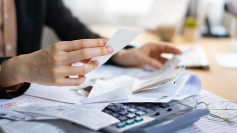 A woman's hands leaf through bills over a table with a calculator underneath.