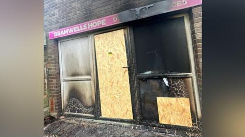 The front of a shop which has been badly damaged by fire. It has a pink sign which says Bramwells Hope and the door and one lower window have been boarded up. 