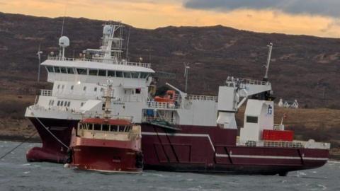 The cargo ship is white and red in colour while the smaller support vessel is similar in colour. The two boats are seen against the background of a rugged hillside.