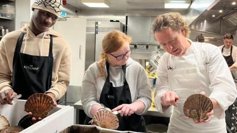 Chef Tom Kitchin, a curly-haired man in chef's whites, demonstrates opening the shell to a young man and woman in his kitchen. They wear dark aprons over their clothes and sous chefs work away in the background
