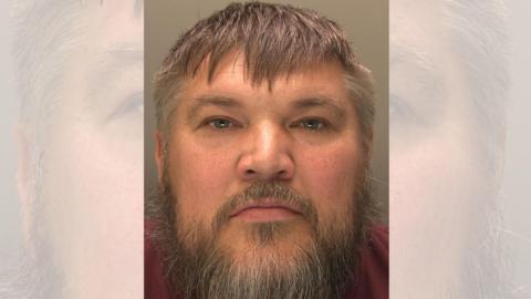 A police custody photo of a man's face taken from close range. He has short, greying brown hair and a greying beard.