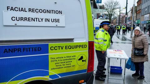 A Thames Valley Police community support officer pictured standing at a table with a woman dressed in a thick coat and woolly hat looking at the camera, with a Thames Valley Police van on the left.
