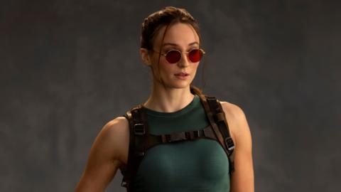 A photo shows Sophie Turner as Lara Croft. She wears red-tinted sunglasses, her hair tied back. She wears a green vest top and tight, thigh-length shorts. One of her fingerless-gloved hands rests on a pistol in a thigh holster.