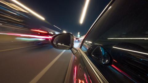 A grey car is speeding along a road at high speed with lights flashing around the car.