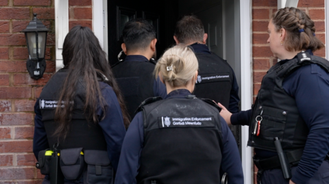 The backs of heads of five immigration enforcement officers who are going into a house. Two are the front are men, and the rest are three women.