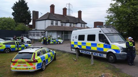 Multiple police cars and a van parked outside the grey hotel building, which is protected by fences. Several officers are standing around on watch.