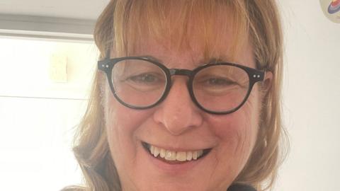 Adele Langlois smiles at the camera while taking a selfie. She has black rimmed glasses on. The top row of her teeth can be seen. She has shoulder length blonde hair.