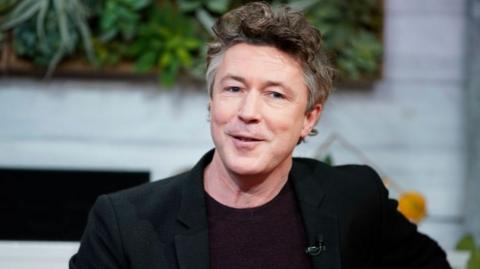 Aidan Gillen looks into the camera. He has brown scruffy hair with some white at the side. He is wearing a black jacket and dark round neck jumper.