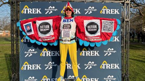 A man dressed in a parrot costume, standing on a small podium in front of a navy blue backdrop at a running race. The costume has red wings with blue feathers on their ends, and a hood that is red, with white around the eyes and a yellow beak. His trousers are yellow. Inside the wings, which are spread, there are two large logos for Action Duchenne. On the right is a black and white QR code and on the left is a photo of a young boy