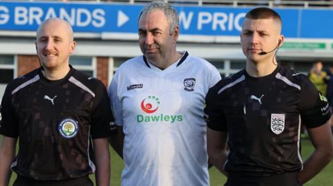 A man with grey-white hair wearing a white Hereford FC shirt stands on a football pitch. On either side of him is a match official wearing black kits and headsets.