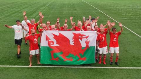 A football team featuring players all over the age of 70. They are holding a Welsh flag with their arms in the air celebrating victory. They are stood on a football pitch with the lines visible. Some have football bots on whereas others have socks or are barefoot.