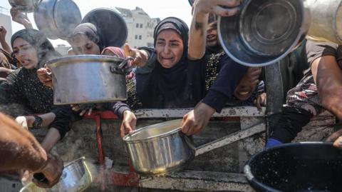 Several people holding pans to fill with food at an aid distribution site - with one woman in the middle wearing all black visibly upset and crying