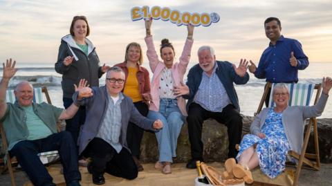 A group of men and women celebrating on a beach with a £1,000,000 sign