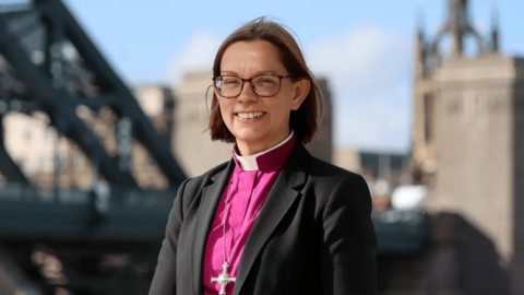 Bishop of Newcastle Helen-Ann Hartley smiles at the camera as she stands in front of the Tyne Bridge. She is wearing a cross and clerical collar on top of her pink shirt and black blazer.