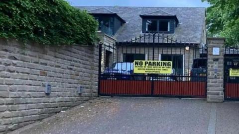 A house pictured from behind some black iron gates. A yellow 'no parking' sign is on the gates.