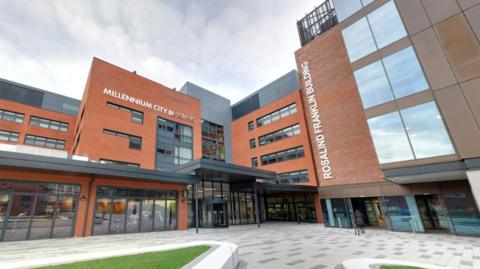 The outside of the University of Wolverhampton's Millennium City and Rosalind Franklin Buildings, both modern multi-storey buildings with glass windows at street level. A grey concrete square with patches of landscaped grass lies outside.