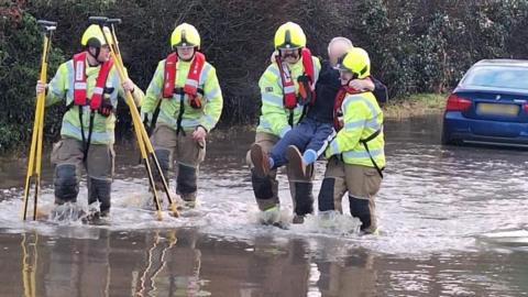 Firefighters in deep water and a blue car parked further down. Two of the firefighters are carrying the elderly man out of the water.