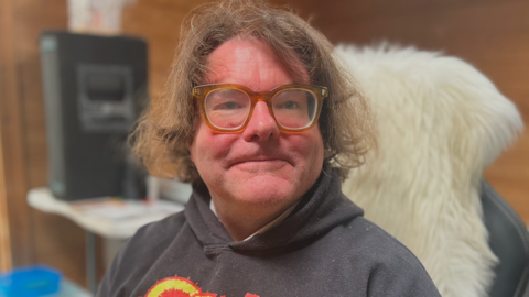 The image shows a man seated indoors, wearing a dark hoodie with a bold graphic on the front and translucent framed glasses. Behind the person is a wooden wall, a large black speaker and a white desk or shelf. He is sitting on a chair with a white, fluffy covering.