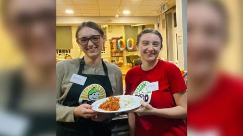 Two women, one wearing a red branded T-shirt  and the other wearing a black branded apron, smiling at the camera and holding up a plate of food. They're standing inside a building and there are balloons saying the number 100 behind them.