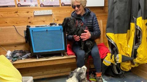A woman sits on a bench next to a heater in a lifeboat rescue station. She has a small black dog on her knee, and three Border Collie type dogs sit at the floor by her feet.