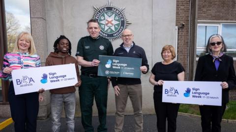 The group are standing in front of a building with the PSNI logo on the wall. They are holding signs promoting Sign Language Week.