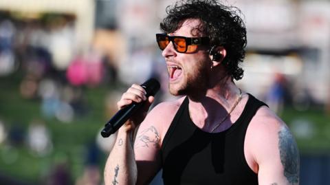Tom Grennan, wearing orange-tinted black glasses and a black vest top, sings into a microphone at Radio 1's Big Weekend in 2023