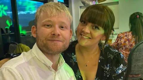 Two people sit close together in a lively indoor setting, one with light blonde hair in a white shirt and the other with brown hair in a dark floral dress, both smiling toward the camera with colourful lights and other guests in the background.