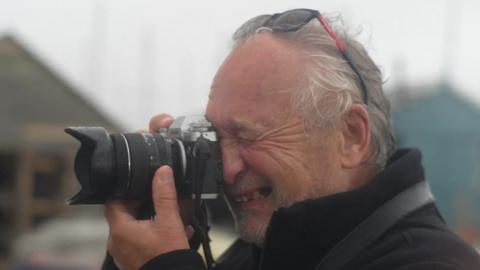 Stephen Squirrell looks into the viewfinder of his camera as he points it to the left. His eye closest to the camera is shut closed as he takes the picture. He has sunglasses on his head and has grey hair, and wears a black jumper. 