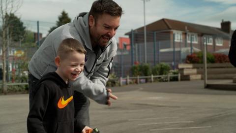 TV presenter Alex Brooker laughing with a school student outside in a school playground.