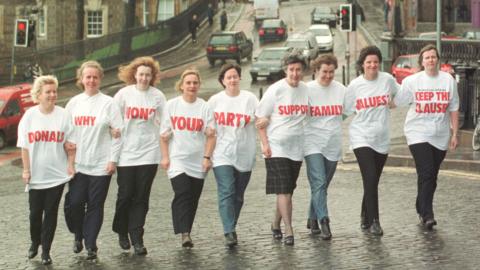 A group of mothers from Perth walk in one long line  to the entrance of the Scottish Parliament - they each wear a white t-shirt with red words which all spell out "Donald, why don't your party support family values, keep the clause".