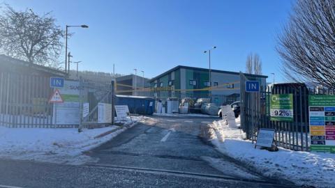 The entrance to a recycling facility is shown on a snowy day, with metal fencing, “IN” signs on both sides of the gate, and several information boards. A green sign promotes local recycling, while modern green‑and‑grey buildings stand behind the fenced area under a clear blue sky.