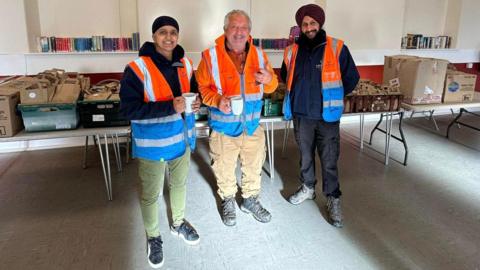 Kully Kaur Deol, Marcus Lapsa and Bill Mato are standing in front of a table laden with sandwich meal bags and boxes of crisps in the Trallwn Community Centre in Pontypridd. They are all smiling and Kully Kaur Deol and Marcus Lapsa are holding cups, with Marcus Lapsa holding what looks like a mobile phone.