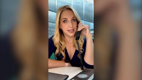 A vertical image of a woman with blonde wavy hair, sitting at a desk and holding a telephone