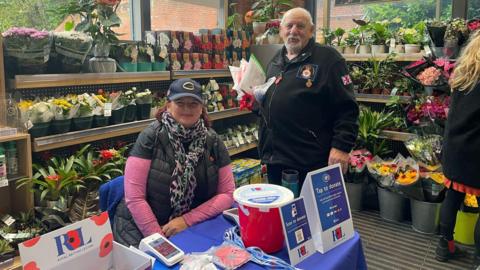 Karen Pitney is sitting by a table with a blue tablecloth selling poppy pins. She is in a garden centre with flowers around her. 