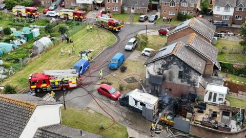 Scorched side of house with firefighter dousing blackened area, gold car and camper van