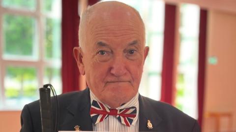 A head-and-shoulders shot of a man wearing a union flag bow tie and a navy suit. He has a couple of gold badges pinned to the jacket. In his hand, he is holding a White Cane with a black handle..