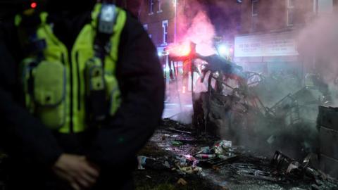 Smoke rises from a burnt out bus in the background with a police officer in the foreground