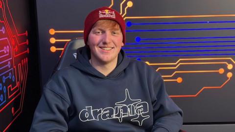 A man sits smiling at the camera in a blue hoodie and a red beanie hat with the words Red Bull on it. He has red hair and is in a room with colourful lines on the walls