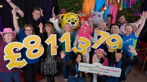 A group of people are celebrating and smiling with their arms raised in the air. Among them is a large tiger costume and a large hippo costume. Between them, they are holding up large yellow numbers that create the number £811,379