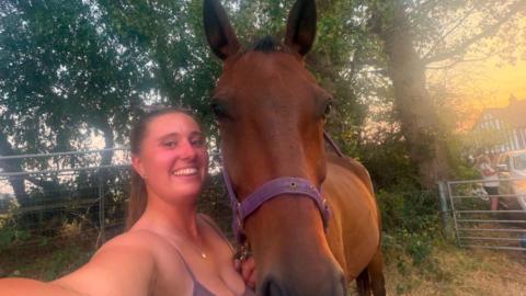 Abbie Harvey takes a selfie while standing next to a brown horse. She has tied back brown hair and is wearing a sleeveless purple top while stood with the horse in a field.