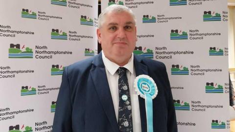 Lee Duffy standing in front of a council banner, looking at the camera. 
He is wearing a dark suit, with a floral tie, a white shirt and a Reform UK rosette on his label.