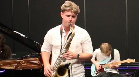 Conor Parker-Delves, playing the saxophone which was stolen in a concert. He has short floppy brown hair and is wearing a white shirt with his sleeves rolled up. In the background is a bass guitar player sitting on a chair, and a person playing a grand piano.