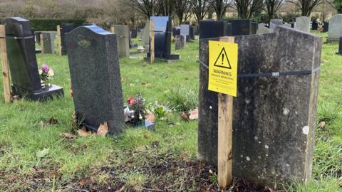 A headstone, with a wooden stick behind it, displaying a yellow 'Warning, Unsafe Material' label