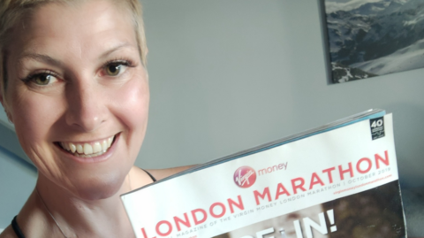A smiley woman with closely cropped blonde hair holds up a magazine with London Marathon printed on the front 