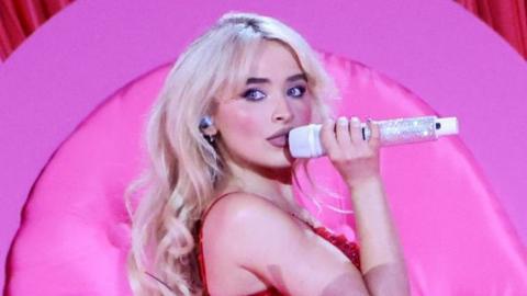 A photo of Sabrina Carpenter performing on stage in front of a pink background holding a sparkling microphone